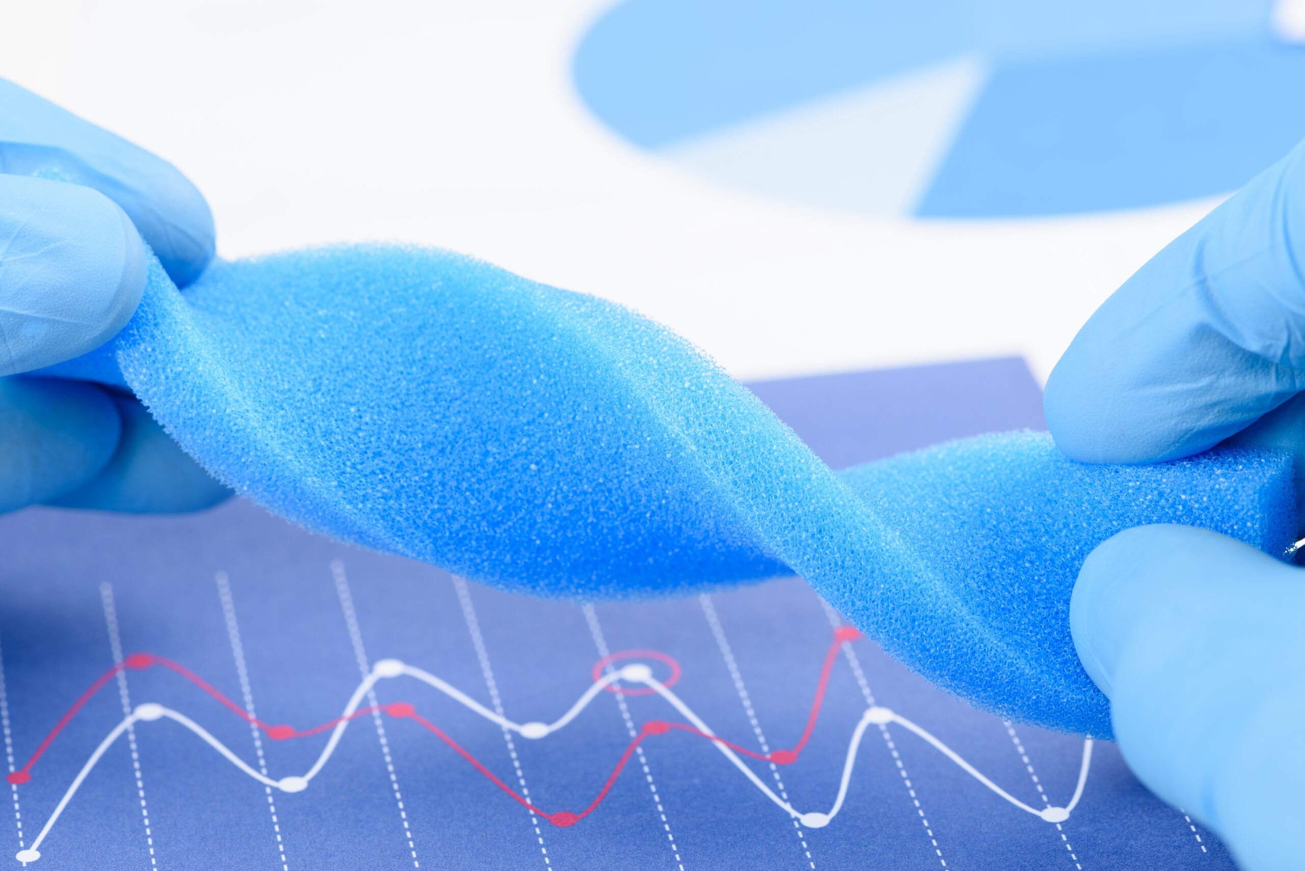 Close-up of gloved hands stretching a blue medical foam material over a chart with performance graphs, illustrating the flexibility, durability, and testing of engineered foam used in medical device applications.